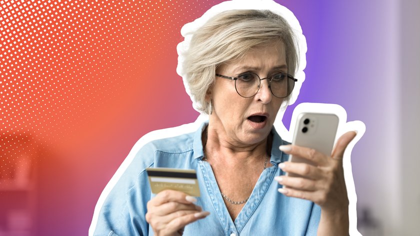 Frau mit weißer Kurzhaarfrisur, Brille und blauem Oberteil schaut schockiert auf ihr Handy, dass sie in der linken Hand hält. In der rechten Hand hat sie eine Kreditkarte. Im Hintergrund ist ein Farbverlauf von Rot zu Blau und ein unscharfer Teil einer Wohnung zu sehen.